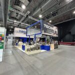 Przestronna przemysłowa hala wystawowa z wysokimi sufitami przedstawia imponujące stoisko targowe Technica Group, prezentujące zbiorniki i rury ze stali nierdzewnej. Dominują niebieskie i białe elementy, z rzędami pustych czerwonych krzeseł po prawej stronie i banerami wiszącymi nad głowami. projekt i nadzór Tatjana Kobuszewska trksystem.com