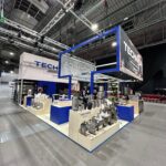 Stoisko targowe Technica Group prezentuje błyszczący przemysłowy sprzęt kuchenny na niebiesko-białych platformach. Nad głową widnieje napis TECHNICA w dużej hali wypełnionej lampami sufitowymi, metalowymi belkami i sąsiednimi kabinami. projekt i nadzór Tatjana Kobuszewska trksystem.com