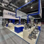 Nowoczesne stoisko targowe firmy Technica Group prezentuje sprzęt kuchenny ze stali nierdzewnej na kremowych i niebieskich ladach pod kwadratową niebieską ramą. Duże oznakowanie TECHNICA znajduje się nad głową, a reflektory oświetlają przestronną salę eventową z wysokimi sufitami. projekt i nadzór Tatjana Kobuszewska trksystem.com