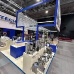 Stoisko targowe prezentuje urządzenia kuchenne i naczynia ze stali nierdzewnej na białych stojakach. Niebieskie ramki z logo Technica Group otaczają stoisko. Wysoki sufit odsłania odsłonięte światła i rury, a w tle pojawiają się rzędy czerwonych krzeseł. projekt i nadzór Tatjana Kobuszewska trksystem.com