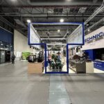 Nowoczesne stoisko targowe Technica Group ma niebieską metalową ramę, wiszące wyraźne znaki TECHNICA i rośliny doniczkowe w centrum. Sprzęt przemysłowy jest prezentowany po prawej stronie w przestronnej hali z wysokimi sufitami i odsłoniętymi belkami. projekt i nadzór Tatjana Kobuszewska trksystem.com