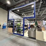 Nowoczesne stoisko targowe Technica Group ma niebieską otwartą prostokątną ramę, drewniane lady, rośliny doniczkowe, urządzenia kuchenne i krzesła. Górne światła rozjaśniają industrialną przestrzeń; przezroczysty znak z odwróconym tekstem wisi w lewym górnym rogu. projekt i nadzór Tatjana Kobuszewska trksystem.com