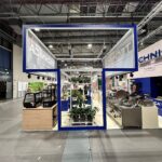 Nowoczesne, otwarte stoisko targowe z przeszklonymi ścianami w niebieskich ramach prezentuje komercyjny sprzęt kuchenny i rośliny. Nazwa Technica Group widnieje na banerach reklamowych. Rzędy lad, maszyn i zieleni są widoczne wewnątrz pod wysokim sufitem z odsłoniętymi belkami. projekt i nadzór Tatjana Kobuszewska trksystem.com