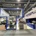 Nowoczesne stoisko targowe prezentuje branding TECHNICA, niebieskie i białe odcienie, industrialny sprzęt kuchenny, zielone rośliny, eleganckie lady ekspozycyjne i metalową ramę nad głową. Dobrze oświetlona przestrzeń ma odsłonięte belki stropowe i reflektory nad nimi. projekt i nadzór Tatjana Kobuszewska trksystem.com
