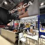 Nowoczesne stoisko targowe dla Technica Group zawiera sprzęt kuchenny na ladach, ekspres do kawy i taborety. Powyżej znajduje się duży czarny baner z napisem "TECHNICA" i "To tworzy profesjonalną kuchnię", ustawiony pośród różnych wyświetlaczy i miejsc do siedzenia. projekt i nadzór Tatjana Kobuszewska trksystem.com