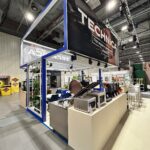 Nowoczesne stoisko targowe z niebieską ramą i dużym napisem "TECHNICA" nad głową prezentuje urządzenia kuchenne Technica Group na eleganckich białych ladach. Przemysłowe lampy sufitowe oświetlają stoisko, które stoi wśród innych żywych ekspozycji wystawowych. projekt i nadzór Tatjana Kobuszewska trksystem.com