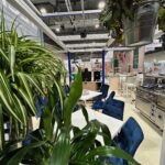 Nowoczesna przestrzeń wewnętrzna z bujnymi zielonymi roślinami na pierwszym planie, niebieskimi wyściełanymi krzesłami wokół białych stołów i urządzeniami kuchennymi ze stali nierdzewnej w tle. Industrialne lampy sufitowe i wiszące rośliny wzbogacają to stylowe stoisko targowe Technica Group. projekt i nadzór Tatjana Kobuszewska trksystem.com