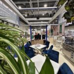 Wewnętrzny showroom Technica Group oferuje nowoczesne urządzenia kuchenne, białe stoły z niebieskimi krzesłami, rośliny liściaste i jasne oświetlenie. Przestronne stoisko targowe obejmuje półki ekspozycyjne, piekarniki, stołki barowe i czytelne znaki informacyjne. projekt i nadzór Tatjana Kobuszewska trksystem.com