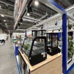 Nowoczesne stoisko targowe autorstwa Technica Group obejmuje szklane gabloty na drewnianej ladzie, prezentujące pustą ekspozycję żywności w hali wystawienniczej. Niebieskie ramy, rośliny i oznakowanie uzupełniają wygląd, z innymi stoiskami i oświetleniem przemysłowym w zasięgu wzroku. projekt i nadzór Tatjana Kobuszewska trksystem.com