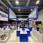 Stoisko targowe Technica Group prezentuje stalowe wyposażenie kuchni: garnki, patelnie i bemary. Nad białymi ladami wiszą niebieskie szyldy TECHNICA. W tle widoczne są oświetlenie, miejsca siedzące i inne stoiska wystawowe. projekt i nadzór Tatjana Kobuszewska trksystem.com