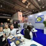 Stoisko targowe z niebieskim dywanem przedstawia ludzi wchodzących w interakcje, broszury i magazyny żywnościowe na ladzie, rośliny doniczkowe i zdjęcie żywności na tylnej ścianie. Górne światła oświetlają Délifrance, Technica Group i inne banery w tętniącej życiem hali. projekt i nadzór Tatjana Kobuszewska trksystem.com