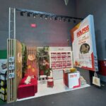 Stoisko targowe firmy DOLFOS ma czerwono-białe kolory, ekspozycję produktów z suplementami na półkach, czerwoną sofę, rośliny doniczkowe i ladę z oznakowaniem marki. Duży baner wyświetla zdjęcia produktów i polski tekst "Sprawność na Maxa". projekt i nadzór Tatjana Kobuszewska trksystem.com