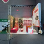 Stoisko modułowe dla Dolfos zawiera czerwono-biały branding, półki z butelkami produktów, ladę i plakaty. Stoisko ozdobione jest wiszącymi owocami i zielenią, a także częścią wypoczynkową z czerwoną ławką. Sąsiednie stoiska są częściowo widoczne. projekt i nadzór Tatjana Kobuszewska trksystem.com