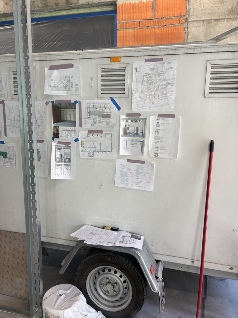 Biała ściana przyczepy przedstawia różne plany i schematy konstrukcyjne, niektóre dla nadchodzącej restauracji chińskiej Halo Mian, przyklejone taśmą w uporządkowany sposób. Papiery spoczywają na małej przyczepie, a czerwona miotła opiera się o ścianę i pojawia się ceglane tło. projekt i nadzór Tatjana Kobuszewska trksystem.com