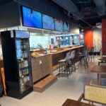 Nowoczesna kawiarnia Halo Mian wyposażona jest w wysoką ladę, szare stołki barowe i czarną lodówkę z napojami. Ekrany wyświetlają treści cyfrowe, a ciepłe oświetlenie i drewniane meble tworzą przytulną atmosferę - idealną do korzystania z tej wyjątkowej restauracji chińskiej. projekt i nadzór Tatjana Kobuszewska trksystem.com