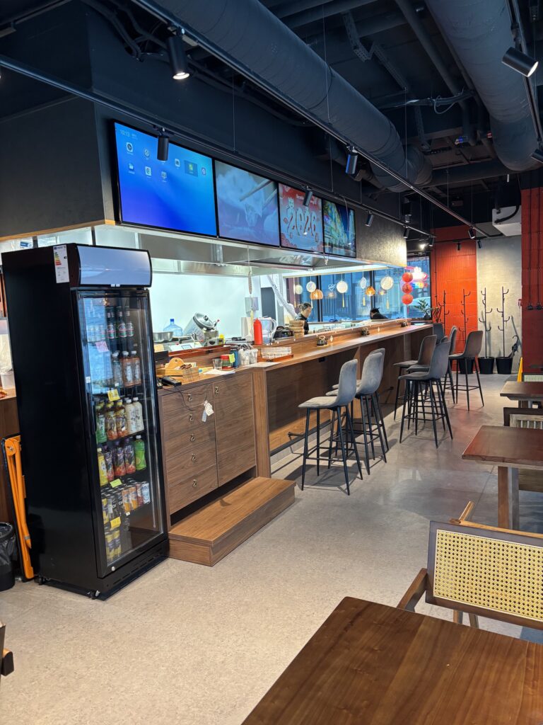 Nowoczesna kawiarnia Halo Mian wyposażona jest w wysoką ladę, szare stołki barowe i czarną lodówkę z napojami. Ekrany wyświetlają treści cyfrowe, a ciepłe oświetlenie i drewniane meble tworzą przytulną atmosferę - idealną do korzystania z tej wyjątkowej restauracji chińskiej. projekt i nadzór Tatjana Kobuszewska trksystem.com