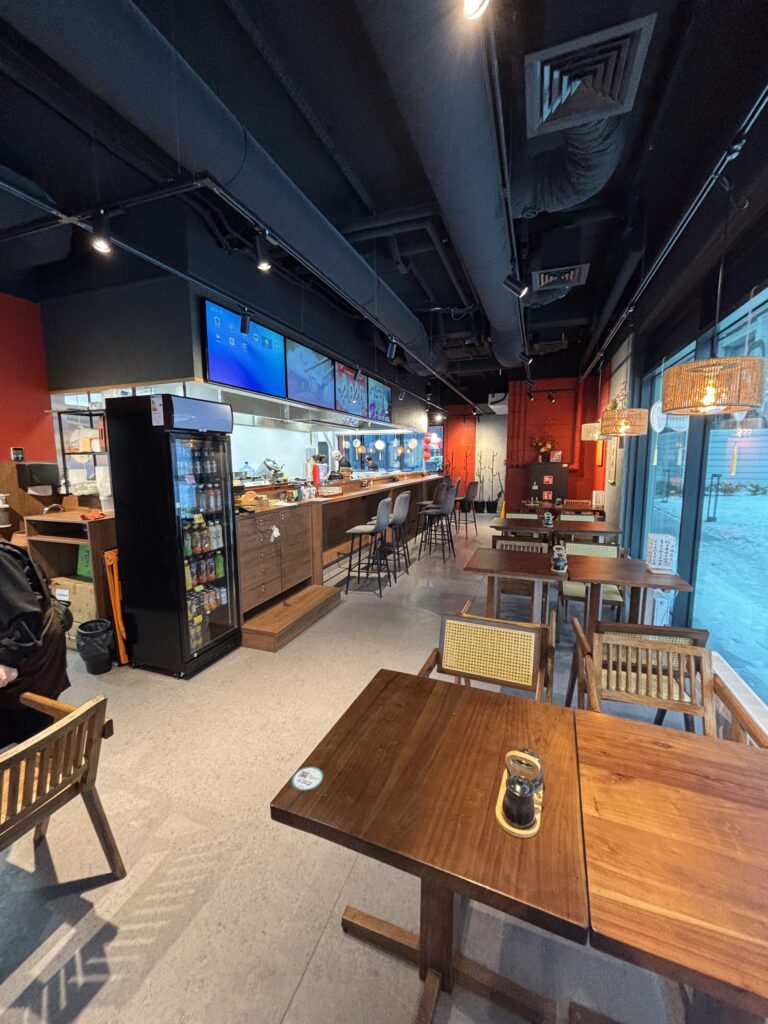 Nowoczesne wnętrze kawiarni z drewnianymi stołami i krzesłami, lampami wiszącymi, ladą ze stołkami i cyfrowym menu nad ladą. Przestrzeń inspirowana Halo Mian restauracja chińska ma czerwono-czarne ściany, duże okna i przytulną atmosferę. projekt i nadzór Tatjana Kobuszewska trksystem.com