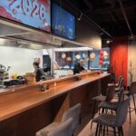 Nowoczesna restauracja chińska z długą drewnianą ladą, szarymi stołkami barowymi i otwartą kuchnią, w której dwóch kucharzy przygotowuje kuchnię chińską. Czerwone i złote lampiony wiszą w oknie, cyfrowe ekrany pokazują rok 2026, a wieszak na płaszcze znajduje się przy czerwonej ścianie. projekt i nadzór Tatjana Kobuszewska trksystem.com