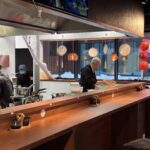 Trzy osoby pracują za drewnianą ladą w Halo Mian, przytulnej restauracji chińskiej. Para unosi się z garnków, bambusowe parowary są ułożone wysoko, a czerwone latarnie świecą przy oknach, podczas gdy na zewnątrz pada śnieg, a czajniki spoczywają na ladzie. projekt i nadzór Tatjana Kobuszewska trksystem.com