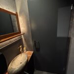 Nowoczesna łazienka w Halo Mian wyposażona jest w kamienną umywalkę na drewnianym blacie, dozownik mydła i okrągłe lustro. Tapeta z motywem natury dodaje uroku, a delikatne oświetlenie dopełnia wygląd - odzwierciedlając zachęcający styl tej restauracji chińskiej. projekt i nadzór Tatjana Kobuszewska trksystem.com