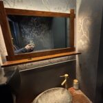 Mała łazienka w Halo Mian, restauracji chińskiej, posiada dekoracyjną kamienną umywalkę na drewnianym blacie, mosiężną baterię i dozownik mydła. Lustro w drewnianej ramie odbija osobę robiącą zdjęcie na tle wzorzystej tapety w gałązki drzew. projekt i nadzór Tatjana Kobuszewska trksystem.com