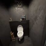 Mała, nowoczesna łazienka z ciemnymi, wzorzystymi ścianami i podłogą - przypominająca stylową restaurację Halo Mian. Biała toaleta znajduje się pośrodku pod półką z dyfuzorami trzcinowymi, bambusowym uchwytem, szczotką i dekoracyjną rzeźbą. projekt i nadzór Tatjana Kobuszewska trksystem.com