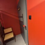 Szare drzwi z czarnym szyldem są osadzone w jasnoczerwonej ścianie restauracji Halo Mian. Obok drzwi stoi nowoczesne krzesło z drewnianą ramą i plecionym siedziskiem, a przy klamce zamontowano czarny włącznik światła. projekt i nadzór Tatjana Kobuszewska trksystem.com