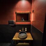 Osoba robi sobie lustrzane selfie w słabo oświetlonej łazience w restauracji chińskiej Halo Mian, z czerwonymi ścianami z geometrycznymi wzorami. Poniżej lustra znajduje się drewniana toaletka z umywalką, dozownikami mydła i podajnikiem ręczników papierowych na lewej ścianie. projekt i nadzór Tatjana Kobuszewska trksystem.com