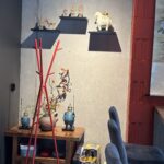 W nowoczesnym pokoju znajdują się dwa szare krzesła, drewniana szafka z wazonem z kwiatami, dwie niebieskie figurki i ułożone w stos pudełka. Z przodu stoi czerwony wieszak na ubrania. W Halo Mian trzy figurki zwierząt i ludzi znajdują się na półkach ściennych nad figurką białego konia. projekt i nadzór Tatjana Kobuszewska trksystem.com