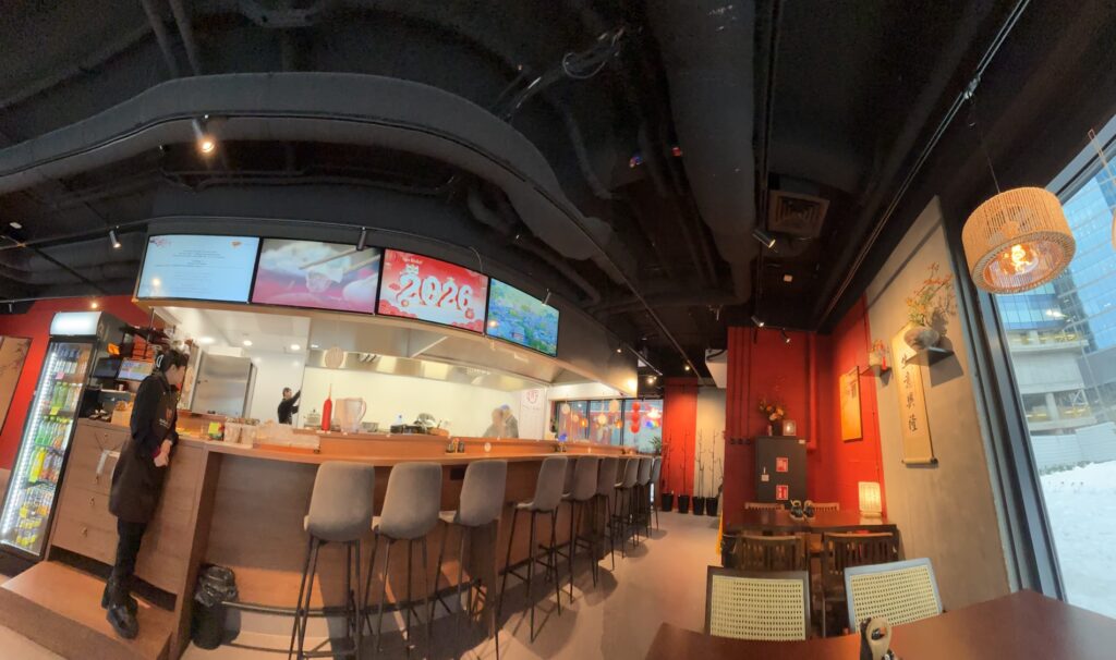 Halo Mian to nowoczesna restauracja chińska z długą drewnianą ladą wyłożoną szarymi krzesłami, pracującym za nią szefem kuchni i podświetlanymi ekranami menu powyżej. Czerwone ściany zdobią azjatyckie dekoracje i obrazy, a duże okna ukazują zaśnieżony krajobraz miasta. projekt i nadzór Tatjana Kobuszewska trksystem.com