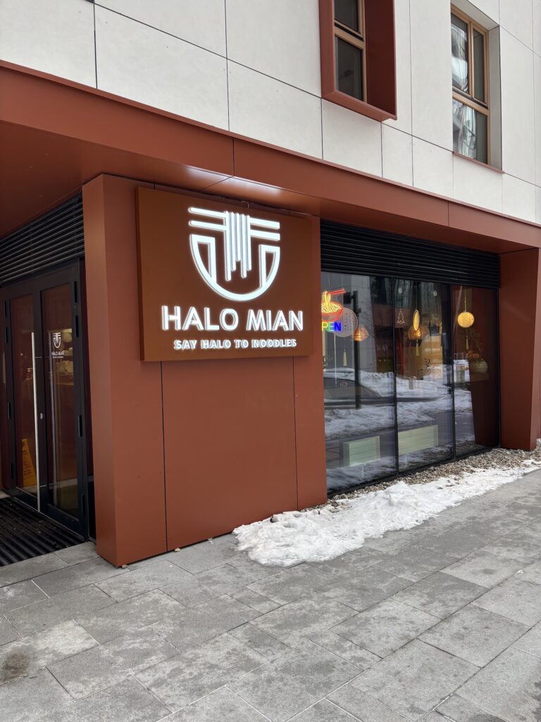 Nowoczesna restauracja chińska z brązowo-białą fasadą. Duży podświetlany napis HALO MIAN nad sloganem SAY HALO TO NOODLES. Na chodniku leży śnieg, a wewnątrz świecą ciepłe światła i neon OPEN. projekt i nadzór Tatjana Kobuszewska trksystem.com