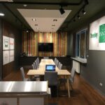 Nowoczesna sala kulinarna z długim drewnianym stołem, szarymi krzesłami, dużym telewizorem i laptopem na stole. Tabliczki z napisami "Dewey Culinary Innovation Studio" i "Develey studio kulinarne". Dobrze oświetlony pokój ma drewnianą podłogę. projekt i nadzór Tatjana Kobuszewska trksystem.com