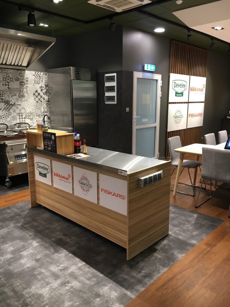 Develey Nowoczesny showroom kuchenny, zainspirowany przez Develey studio kulinarne, obejmuje drewnianą wyspę z markowymi panelami (Develey, Kikkoman, Tabasco, Fiskars), urządzenia ze stali nierdzewnej, wzorzyste płytki, przytulną jadalnię, szare ściany ozdobione sponsorami i drewnianą / dywanową podłogę. projekt i nadzór Tatjana Kobuszewska trksystem.com