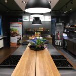 Nowoczesna kuchnia z drewnianymi ladami, czarnymi płytami indukcyjnymi, dużymi wiszącymi czarnymi lampami, półkami z sosami i banerami kulinarnymi Develey studio przedstawiającymi marki spożywcze. Na blacie stoi kosz z sałatą, a na prawej ścianie wiszą garnki i patelnie. projekt i nadzór Tatjana Kobuszewska trksystem.com