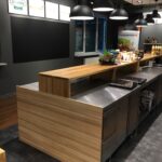 Nowoczesna lada kuchenna z drewnianym wykończeniem i powierzchnią ze stali nierdzewnej stoi pod czarnymi lampami wiszącymi w Develey studio kulinarne. Żywność i warzywa są eksponowane na ladzie, z napisami na ścianie i półkami z produktami w tle. projekt i nadzór Tatjana Kobuszewska trksystem.com