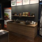 Nowoczesny bufet składa się z drewnianych blatów z dwoma naczyniami do podgrzewania, kubkami i stosem małych talerzy. Powyżej, półka wyświetla ketchup, musztardę i sosy z Develey studio kulinarne. Znaki firmowe zdobią szarą ścianę. projekt i nadzór Tatjana Kobuszewska trksystem.com