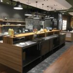 Nowoczesna kuchnia komercyjna w Develey studio kulinarne obejmuje drewniane blaty, urządzenia ze stali nierdzewnej, czarne lampy wiszące, otwarte półki z naczyniami kuchennymi, przyprawy na blacie i jadalnię widoczną w tle - czyste i schludnie zorganizowane. projekt i nadzór Tatjana Kobuszewska trksystem.com