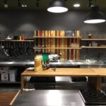 Nowoczesna kuchnia komercyjna autorstwa Develey studio kulinarne obejmuje blaty ze stali nierdzewnej, patelnie wiszące na ścianie, sosy i przyprawy na drewnianym blacie, półki z naczyniami, kuchenkę elektryczną i górne lampy ustawione na ciemnym tle. projekt i nadzór Tatjana Kobuszewska trksystem.com