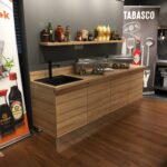 Nowoczesna kuchnia z drewnianymi szafkami, czarnym zlewem i pływającą półką na przyprawy. Banery Kikkoman i Tabasco otaczają kuchnię Develey studio kulinarne. Na blacie stoją metalowe naczynia do smażenia; garnki i patelnie wiszą na ścianie po prawej stronie. projekt i nadzór Tatjana Kobuszewska trksystem.com