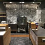 Nowoczesna kuchnia komercyjna z urządzeniami ze stali nierdzewnej, wzorzystymi kafelkami i drewnianymi blatami. Czysta, zorganizowana przestrzeń w Develey studio kulinarne obejmuje piekarnik, lodówkę, napoje butelkowane przy menu lub ulotkach oraz różne narzędzia kuchenne. projekt i nadzór Tatjana Kobuszewska trksystem.com