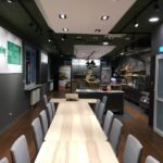 Nowoczesne studio kulinarne Develey ze stołami z jasnego drewna i szarymi krzesłami ustawionymi w rzędach. Pomieszczenie ma ciemne podłogi, zielone i czarne sufity, jasne oświetlenie, sprzęt kuchenny, wyświetlacze żywności i znaki Develey na ścianach. projekt i nadzór Tatjana Kobuszewska trksystem.com