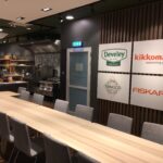 Nowoczesna sala konferencyjna z długim drewnianym stołem i szarymi krzesłami. Za nim znajduje się kuchnia z urządzeniami ze stali nierdzewnej - idealna na sesję kulinarną w studiu Develey. Cztery logotypy marki są wyświetlane na ciemnej ścianie. Na stole stoi laptop. projekt i nadzór Tatjana Kobuszewska trksystem.com