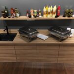 Na podłodze z ciemnego drewna stoi drewniana lada z czarnym zlewem, dwoma przykrytymi naczyniami, białymi talerzami i papierowymi kubkami. Powyżej znajduje się półka z przyprawami i sosami Develey. Konfiguracja tworzy nowoczesny klimat kulinarnego studia Develey na tle szarej ściany. projekt i nadzór Tatjana Kobuszewska trksystem.com