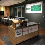 Nowoczesne studio kuchenne z drewnianą ladą recepcyjną prezentującą produkty spożywcze i logo sponsorów, takich jak Develey, Kikkoman, Tabasco i Fiskars; w tle długie stoły z szarymi krzesłami i dużym napisem "Develey studio kulinarne". projekt i nadzór Tatjana Kobuszewska trksystem.com