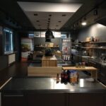 Nowoczesne, słabo oświetlone studio kulinarne Develey z wieloma stanowiskami roboczymi ze stali nierdzewnej, półkami zaopatrzonymi w składniki i przybory kuchenne, banerami reklamującymi marki spożywcze, butelkami i słoikami na ladach oraz ciemnym sufitem z oświetleniem szynowym. projekt i nadzór Tatjana Kobuszewska trksystem.com