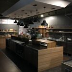 Nowoczesna kuchnia z drewnianymi i metalowymi blatami, wiszące czarne lampy wiszące, otwarte półki z przyprawami i butelkami, przybory i urządzenia na blatach, znak TABAS na tylnej ścianie - to studio Develey jest czyste, zorganizowane i słabo oświetlone. projekt i nadzór Tatjana Kobuszewska trksystem.com