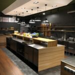 Nowoczesna, przestronna kuchnia autorstwa Develey studio kulinarne charakteryzuje się wykończeniami z drewna i stali nierdzewnej, centralną wyspą, półkami ze słoikami i butelkami oraz eleganckimi urządzeniami. Czarne lampy wiszące wiszą nad głową, oświetlając zorganizowaną, czystą przestrzeń. projekt i nadzór Tatjana Kobuszewska trksystem.com