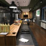 Nowoczesna, dobrze oświetlona kuchnia w Develey studio kulinarne ma długie drewniane blaty, płyty kuchenne, butelki z przyprawami i urządzenia. Stołki barowe ustawione są wzdłuż lady, a jadalnia z białymi krzesłami znajduje się w pobliżu dużych okien z zasłonami w paski. projekt i nadzór Tatjana Kobuszewska trksystem.com