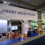Na stoisku targowym znajdują się banery "Henry Kruse Polska" i "bagstar". Przestrzeń jest dobrze oświetlona, prezentując środki czyszczące, produkty przyjazne dla środowiska, stoły, rośliny i siedzenia pod lampami sznurkowymi. Ludzie angażują się w różne stoiska. projekt i nadzór Tatjana Kobuszewska trksystem.com