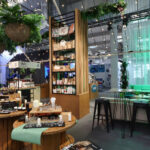 Nowoczesny wewnętrzny market oferuje drewniane stoły i półki z produktami kosmetycznymi, roślinami doniczkowymi i wiszącą zielenią. Czarne stołki ustawione są wzdłuż baru, a delikatne zielone oświetlenie tworzy przytulną, zachęcającą atmosferę z lampkami wróżkowymi nad głową. projekt i nadzór Tatjana Kobuszewska trksystem.com